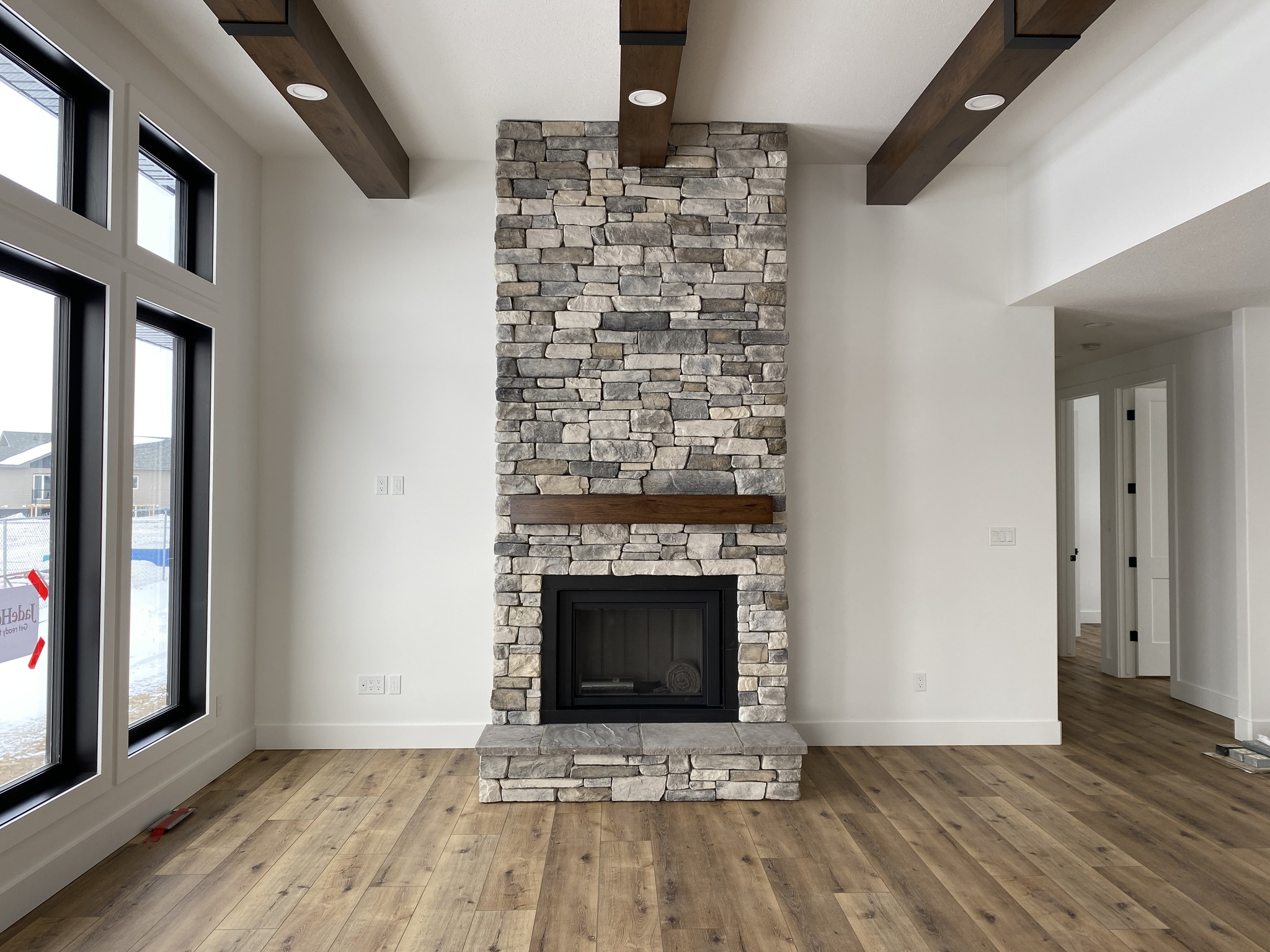 A modern living room with a tall stone fireplace centered against a white wall. Wooden beams adorn the ceiling, and large windows let in natural light.
