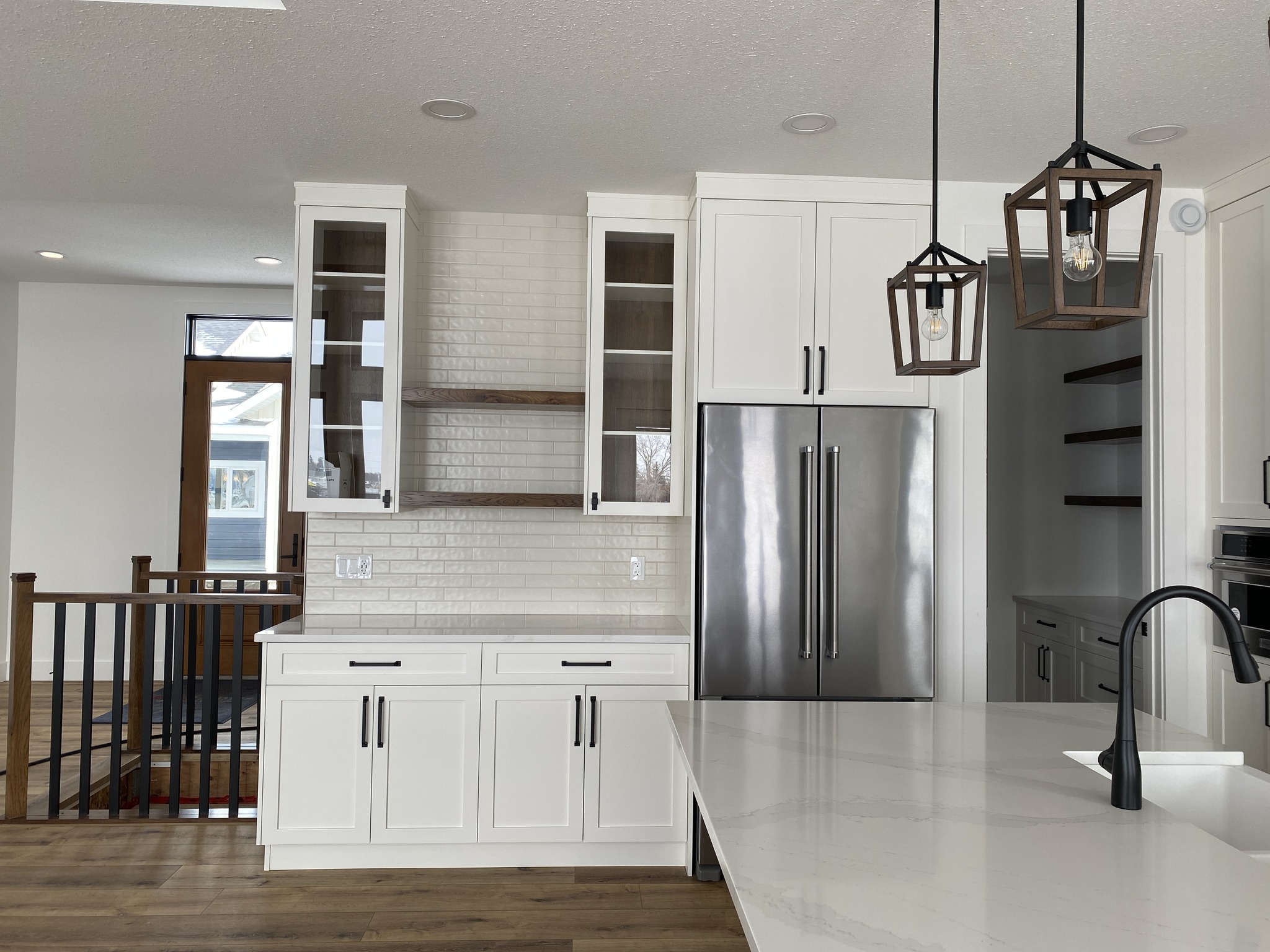 Modern kitchen with white cabinets, stainless steel fridge, and marble countertops. Features pendant lights and wooden floors, creating a sleek, warm ambiance.