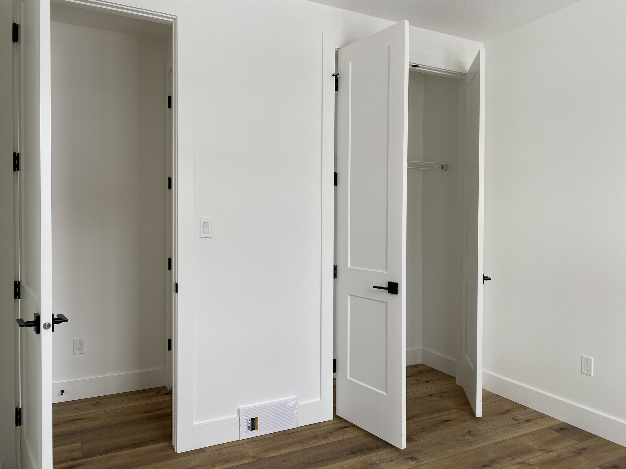 An empty room with wooden flooring and two open white closet doors, revealing empty interiors. The space feels clean, minimalist, and freshly painted.
