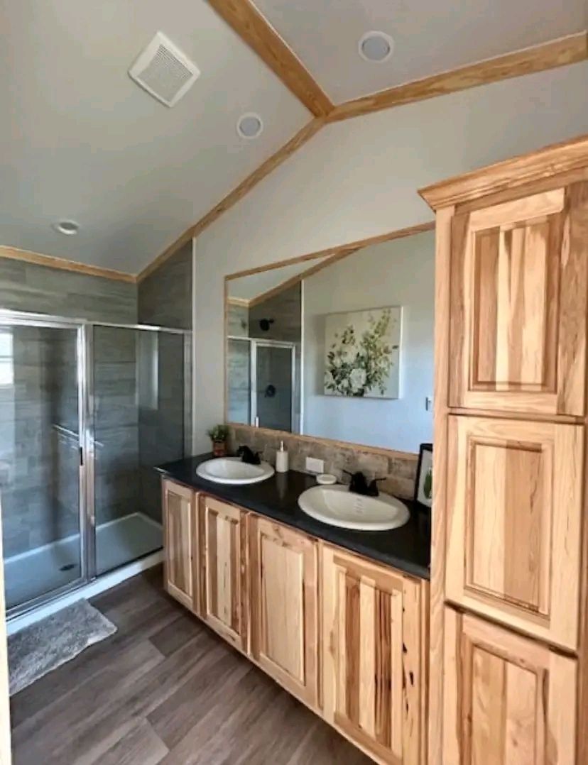 A modern bathroom featuring a double vanity with wooden cabinets, dual sinks, and a large mirror. A glass-enclosed shower and floral artwork add elegance.
