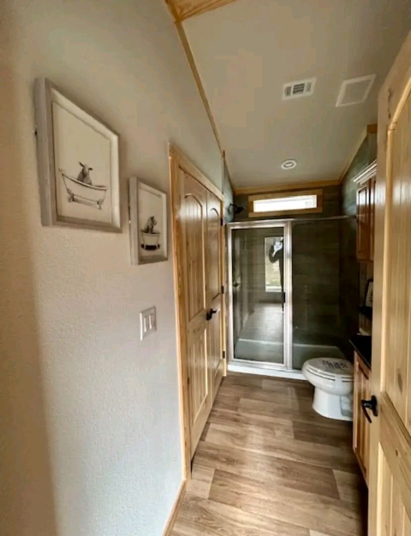 Narrow bathroom with light wood flooring and trim, featuring a shower with glass doors and a white toilet. Two framed boat-themed artworks on the wall. Cozy and rustic feel.