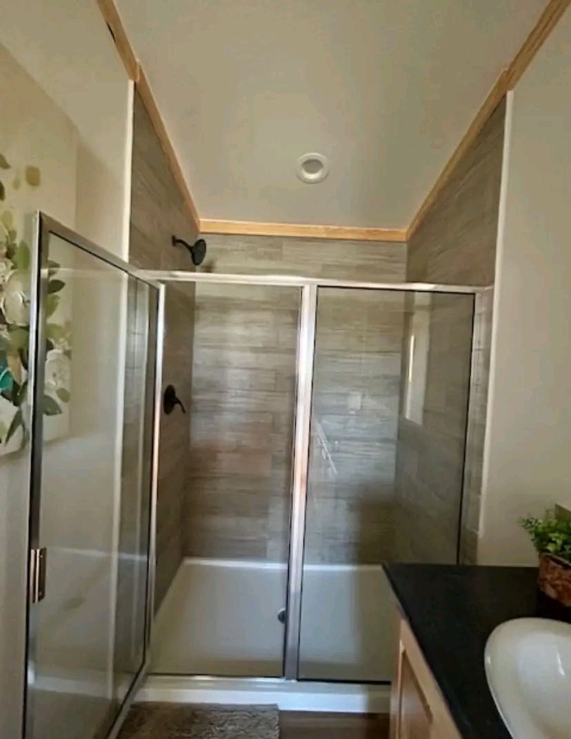Modern bathroom with a glass-enclosed shower featuring gray stone tiles. A small potted plant sits on a dark countertop under soft lighting.