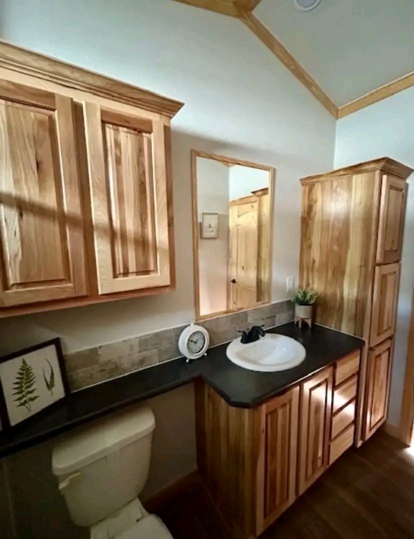A cozy bathroom features natural wood cabinetry, a black countertop with a white sink, a framed mirror, and decorative elements, creating a warm ambiance.