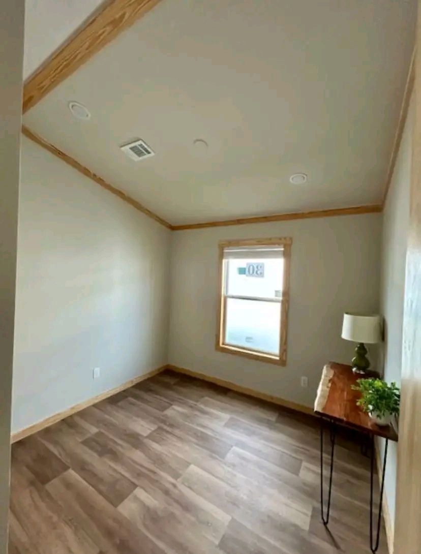 Simple room with light wood flooring and beige walls, featuring a large window. A small wooden table with a lamp and plant adds warmth. Tranquil atmosphere.