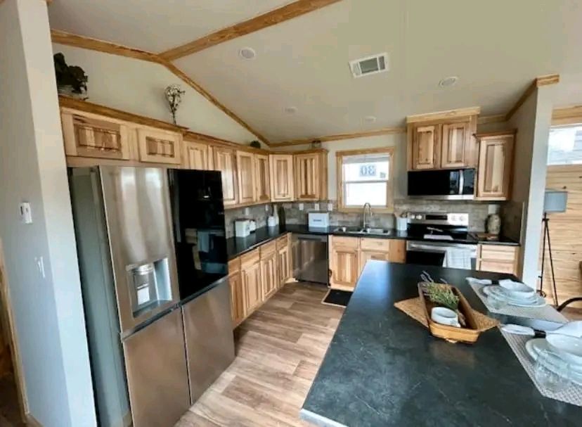 A modern kitchen with wooden cabinets, stainless steel appliances, and a black countertop. The light wood floor and decor create a warm, inviting ambiance.