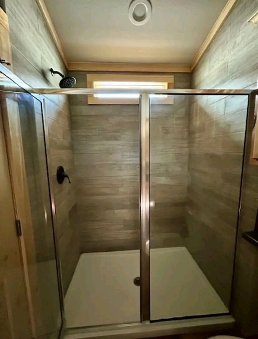 Modern bathroom shower with glass doors, wood-textured tile walls, a small window above, and a mounted black showerhead. Bright and clean ambiance.