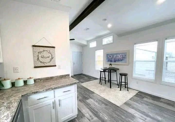 Modern kitchen with white cabinets, marble countertops, and three green jars. Nearby, a small dining area with a round table, two stools, and a wall art piece create a cozy atmosphere. Natural light fills the room.