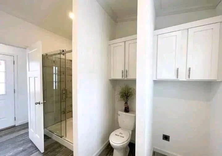 A bathroom with a shower on the left, an open door, and white cabinets above a toilet with a plant. The toilet lid reads “Do Not Use." Bright and tidy.