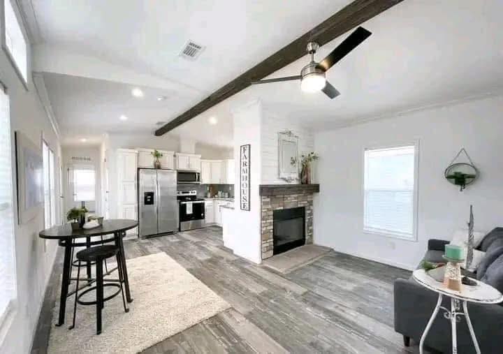 Modern farmhouse interior with an open layout. Features include a rustic wood floor, brick fireplace, ceiling beam, sleek kitchen, and a cozy sofa.