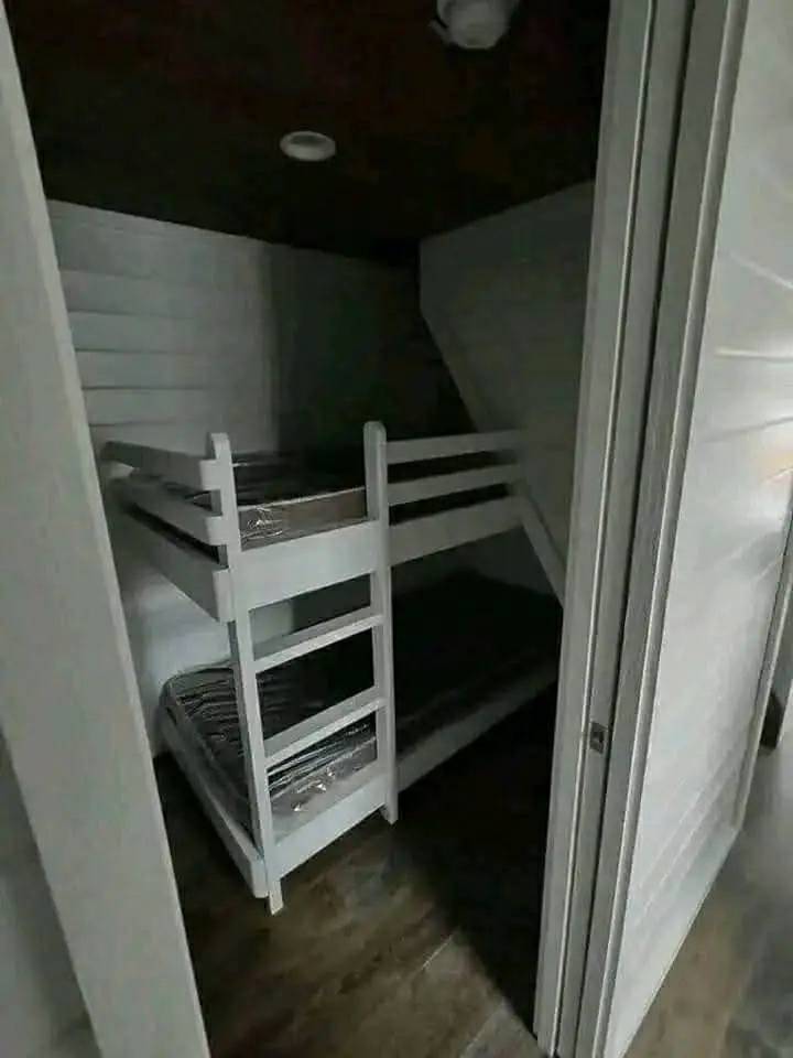 A small, dimly lit room features white wooden bunk beds against white walls and a wooden floor, creating a cozy, minimalist atmosphere.
