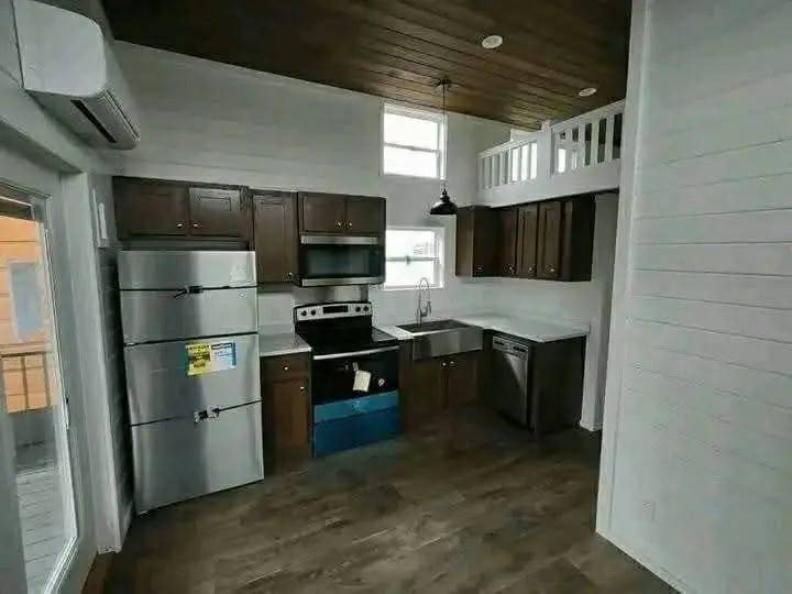 Compact modern kitchen with dark wood cabinets, stainless steel appliances, and a farmhouse sink. High ceiling with loft, wooden floor, and pendant light.
