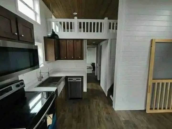 A cozy kitchen with white walls and dark wood cabinets, featuring a stainless steel stove and a black pendant light. A loft area with a white railing is visible above.