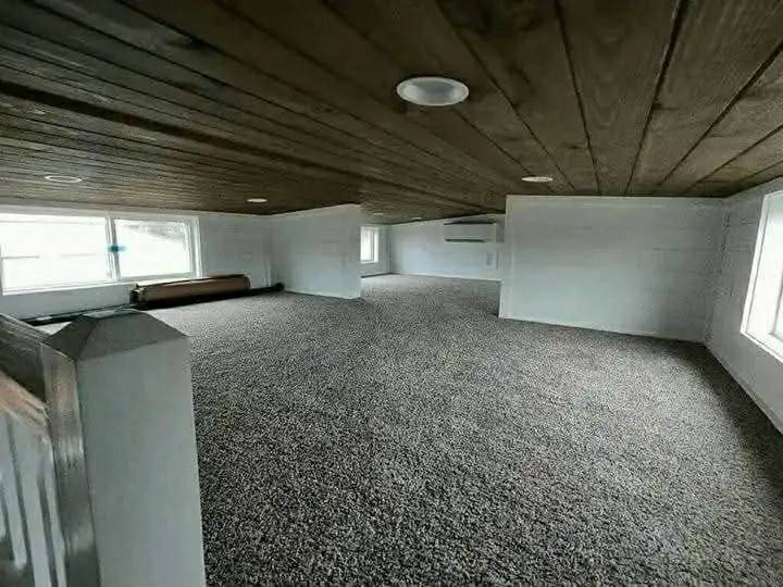 Spacious attic with wooden ceiling, gray carpet, and large windows on both sides, creating a well-lit and cozy atmosphere.