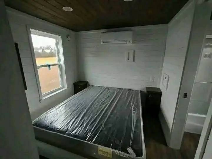 A small bedroom with a queen-sized bed covered in plastic wrap. Two wooden nightstands are on either side. A window lets in natural light, and an AC unit is mounted above the bed, creating a minimalist, cozy atmosphere.