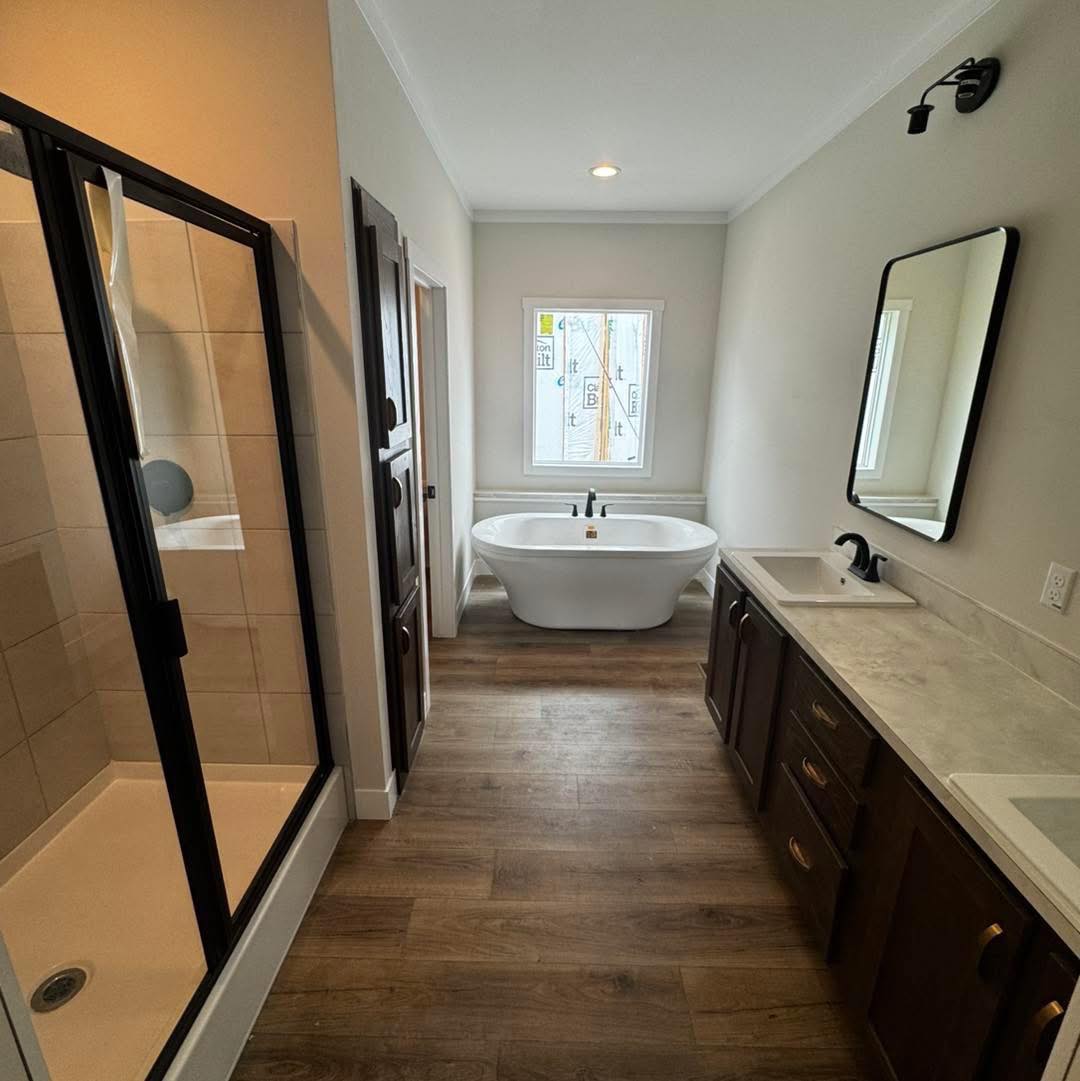 A modern bathroom with a freestanding bathtub under a window, a glass shower on the left, and a dual-sink vanity with dark wood cabinets on the right.