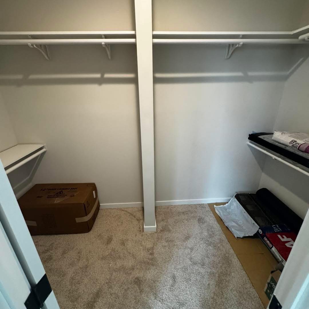 A small, empty walk-in closet with white walls, beige carpet, and shelves under a white rod. A cardboard box sits in the left corner, conveying newness.
