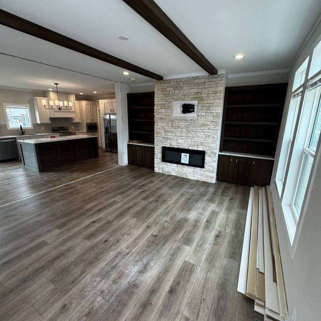 Modern open-concept living room with wood floors, exposed beams, stone fireplace, built-in shelves, and adjacent kitchen with pendant lighting. Cozy and inviting.