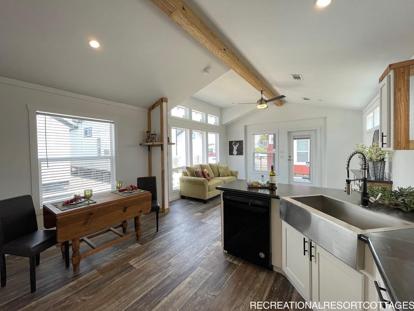 Spacious open-plan living area with wood beam ceiling, large windows, and a cozy sofa. Kitchen features modern fixtures, and a rustic dining table.