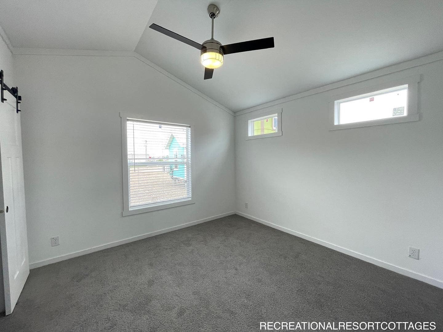 A modern, minimalist room with white walls, gray carpet, and a vaulted ceiling. Features a large window, two small windows, and a ceiling fan.
