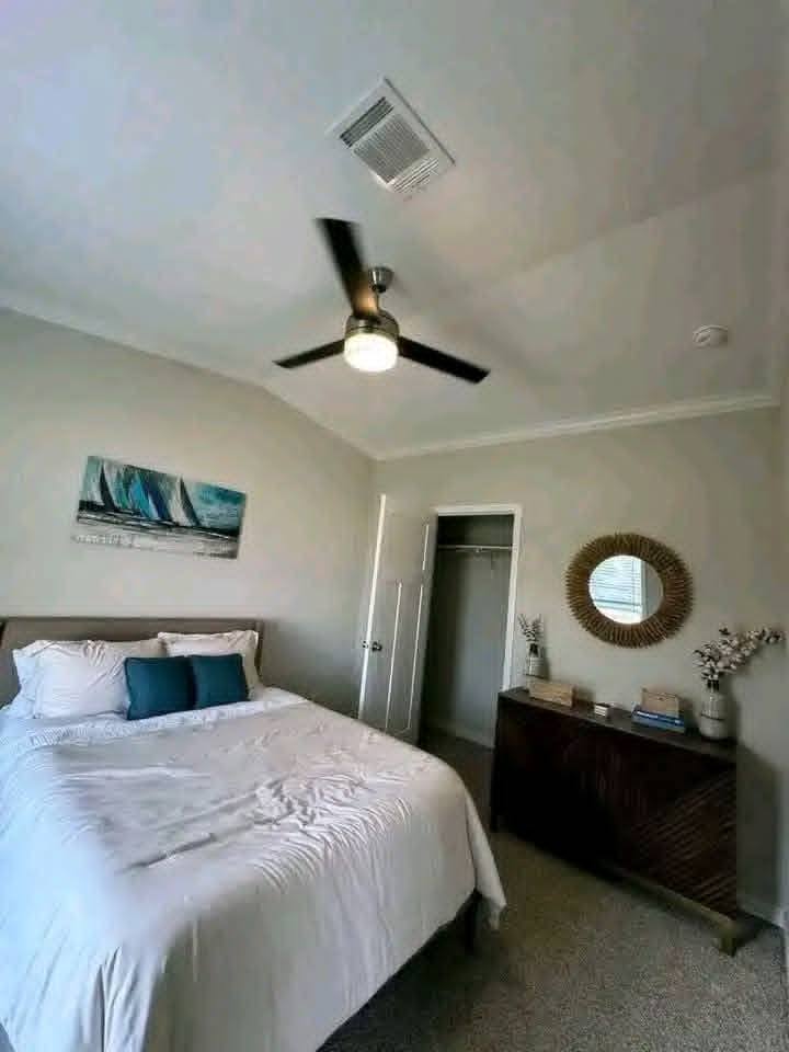 Cozy bedroom with a white bed, teal cushions, and sailboat art. A ceiling fan, closed closet, and dresser with a round mirror add elegance.