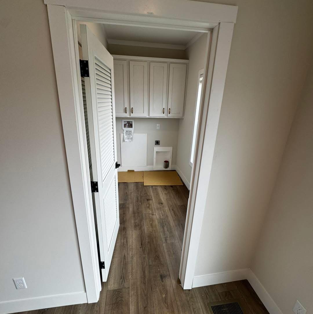 A narrow laundry room with open door, featuring white cabinets, a hardwood floor, and wall cutouts for appliances. Neutral tones create a clean, modern look.