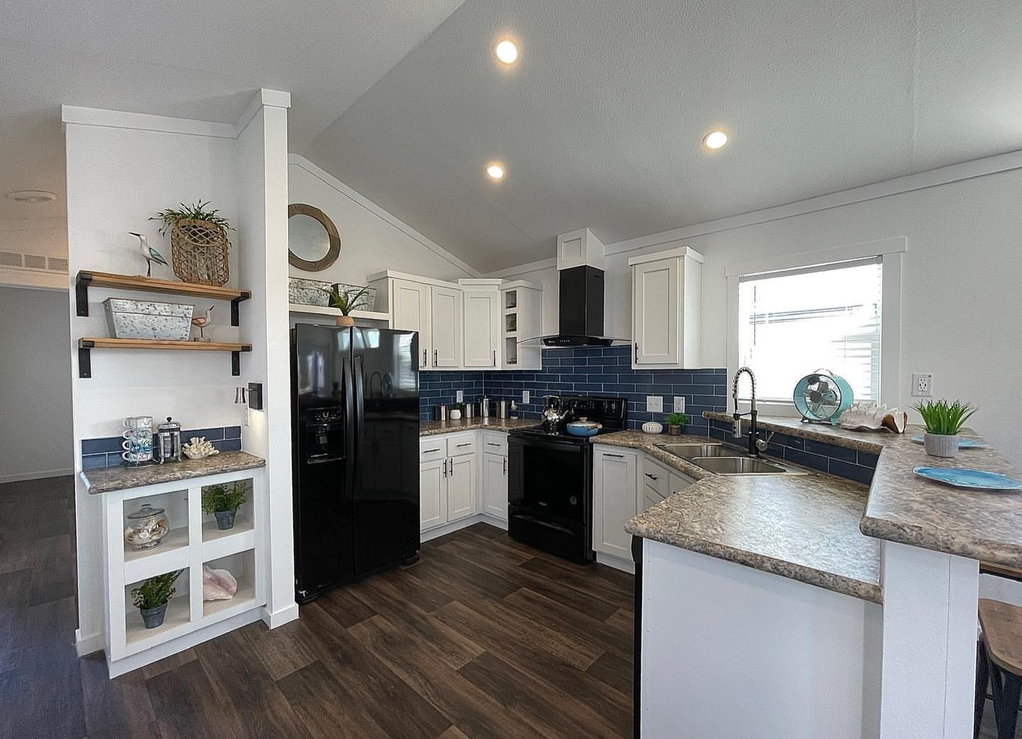 Spacious modern kitchen with white cabinets, dark blue backsplash, and black appliances. Open shelving and plants add warmth. Natural light from a window.
