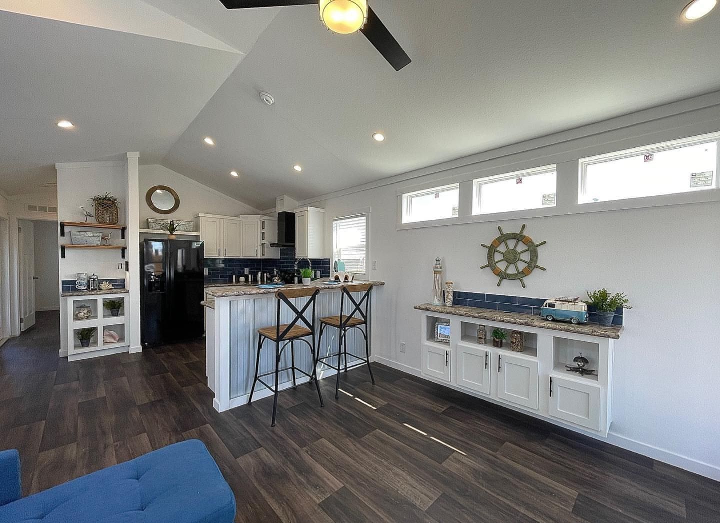 Modern kitchen and living area with dark wood floors, high ceilings, and white walls. Features a bar counter with two chairs, a nautical theme, and decorative shelves.