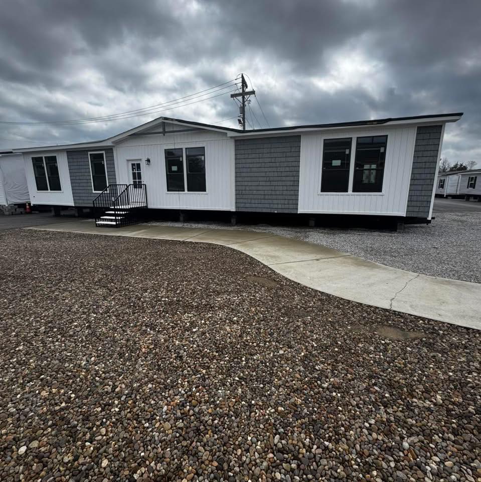 A modern, single-story modular home with gray siding and white accents sits under a cloudy sky. The foreground shows a gravel path and a small concrete walkway.