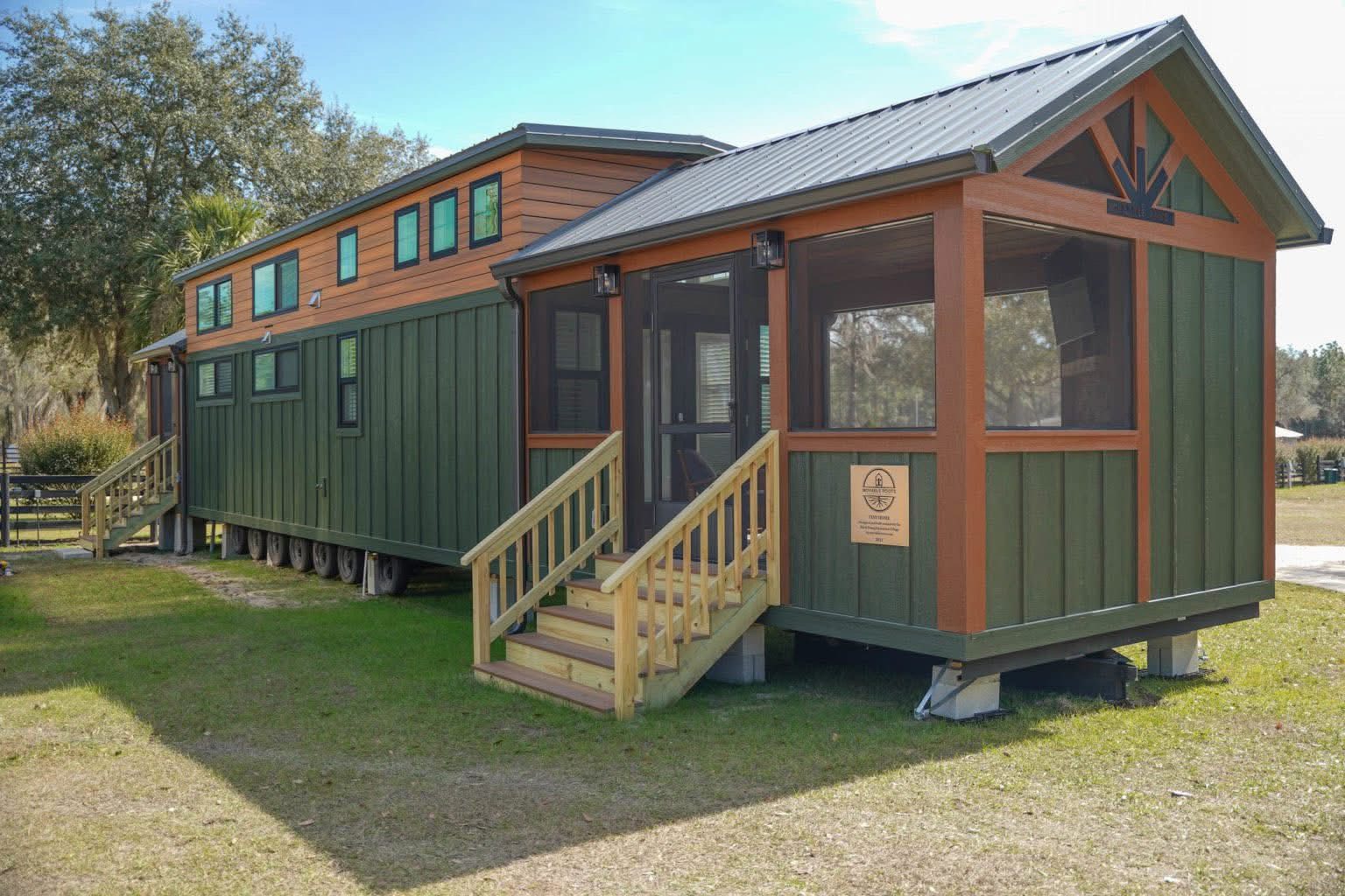 A modern tiny house with green and brown paneling, featuring multiple windows and a screened porch. It stands on a grassy area under a clear blue sky.