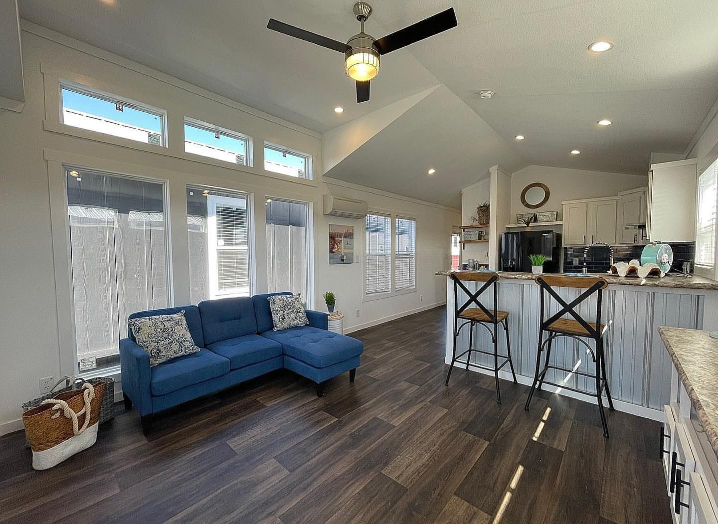 Modern living area with a vaulted ceiling, featuring a blue sectional sofa, large windows, dark wood flooring, and an open kitchen with bar stools. Cozy and bright ambiance.