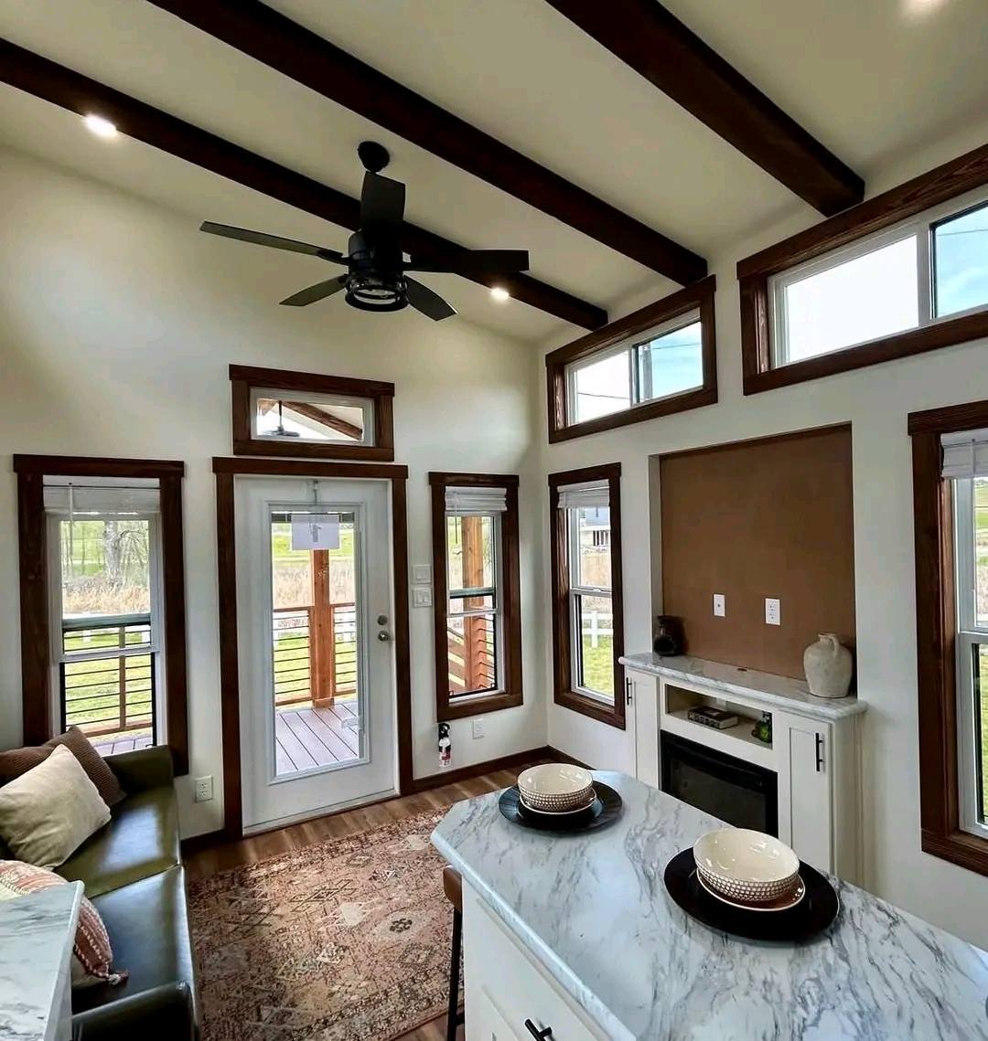 Cozy living area with high ceilings, wooden beams, and a ceiling fan. There's a marble counter, soft couch, fireplace, and bright natural light through large windows.