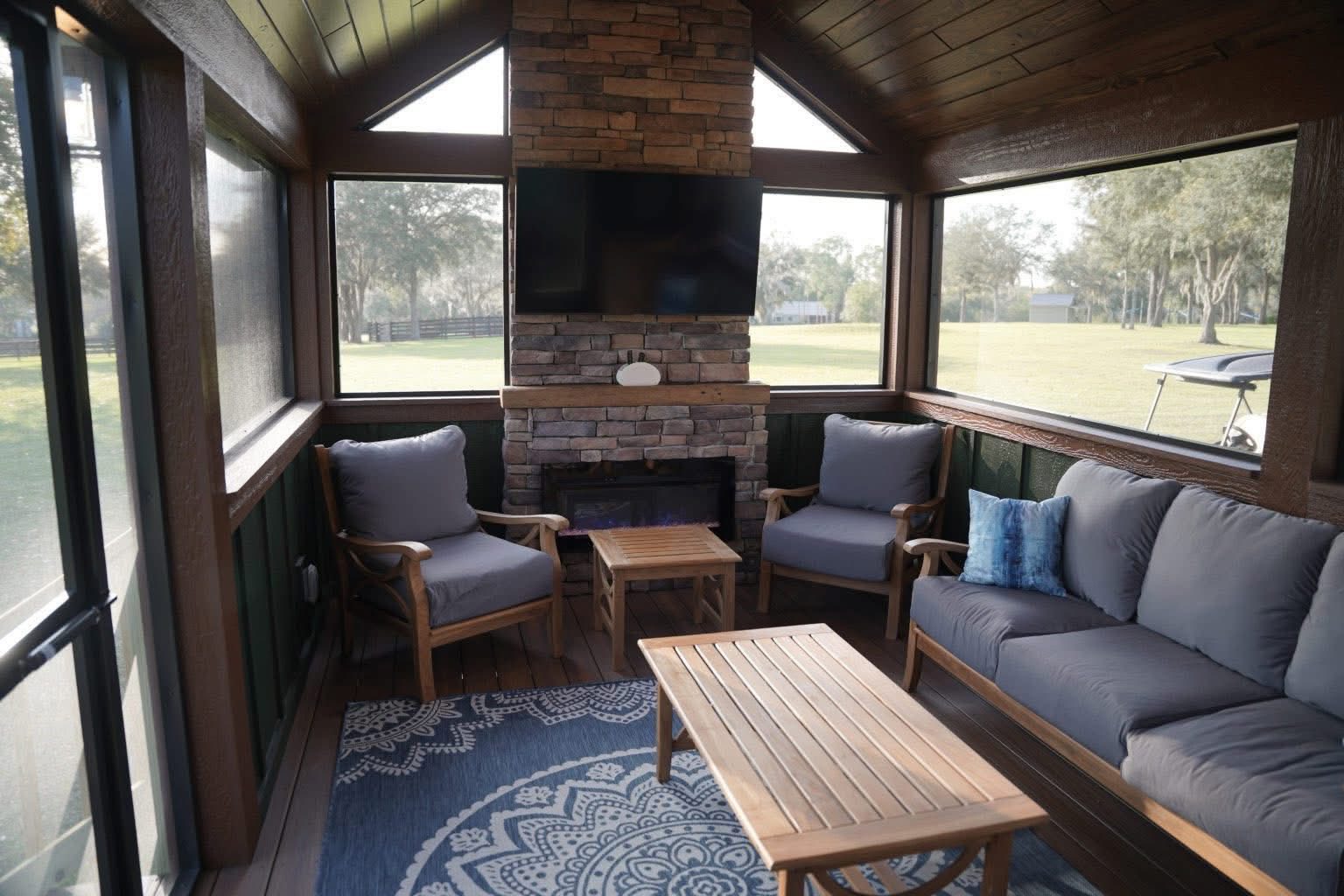 Cozy sunroom with a stone fireplace, TV, and large windows showcasing a grassy field. Features wooden furniture, gray cushions, and a blue patterned rug.