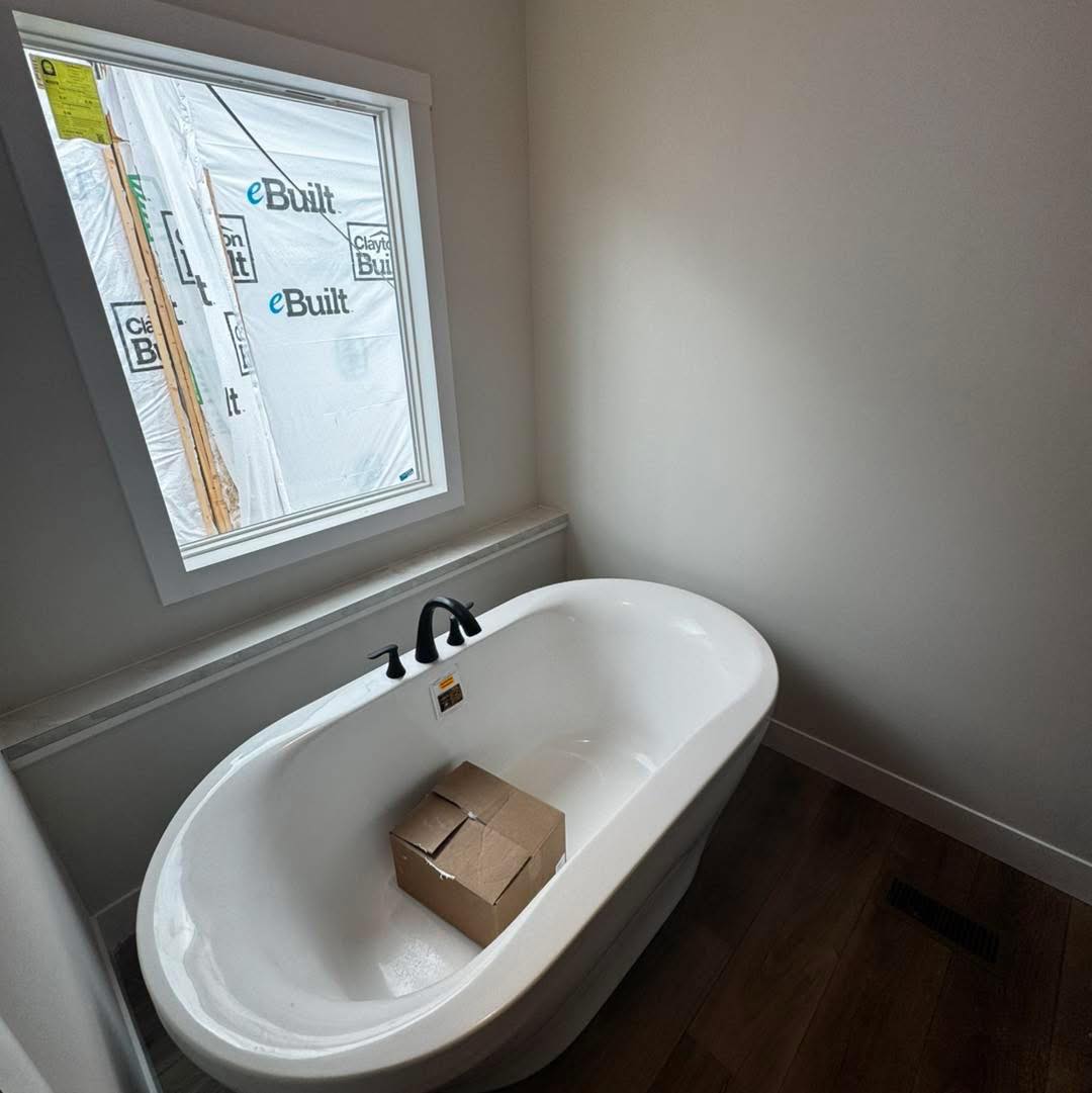 A white bathtub with a cardboard box inside is positioned near a window, through which construction materials are visible. The room has wooden floors and an unfinished feel.
