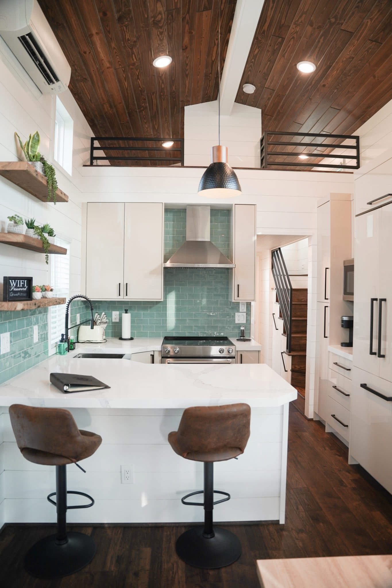 Modern kitchen with white cabinets, teal backsplash, and stainless steel appliances. Two brown stools at a white island, wood ceiling and floors. Cozy ambiance.
