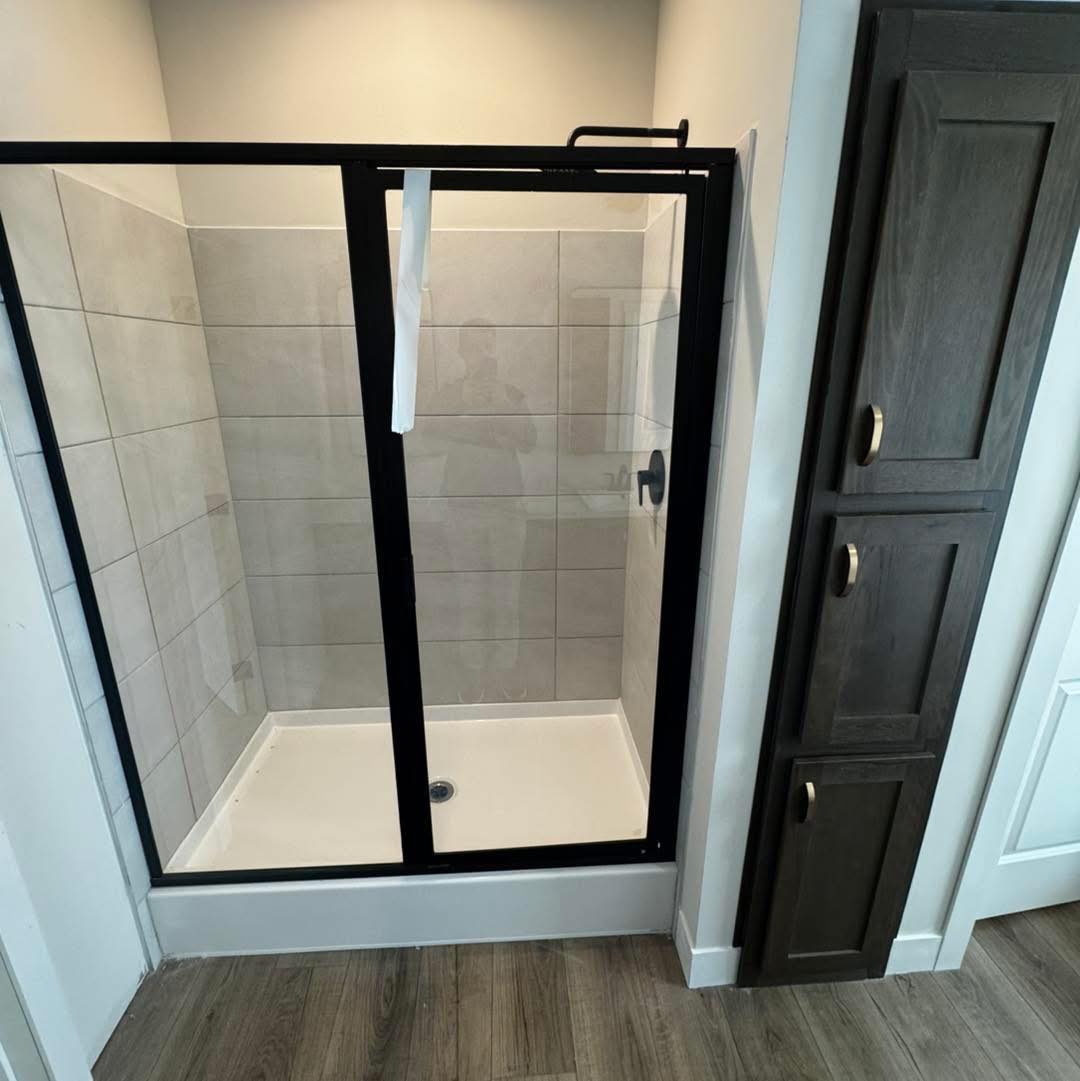 A modern shower with a black metal frame and clear glass door, set against beige tile walls. Next to it, there is a dark wood cabinet.