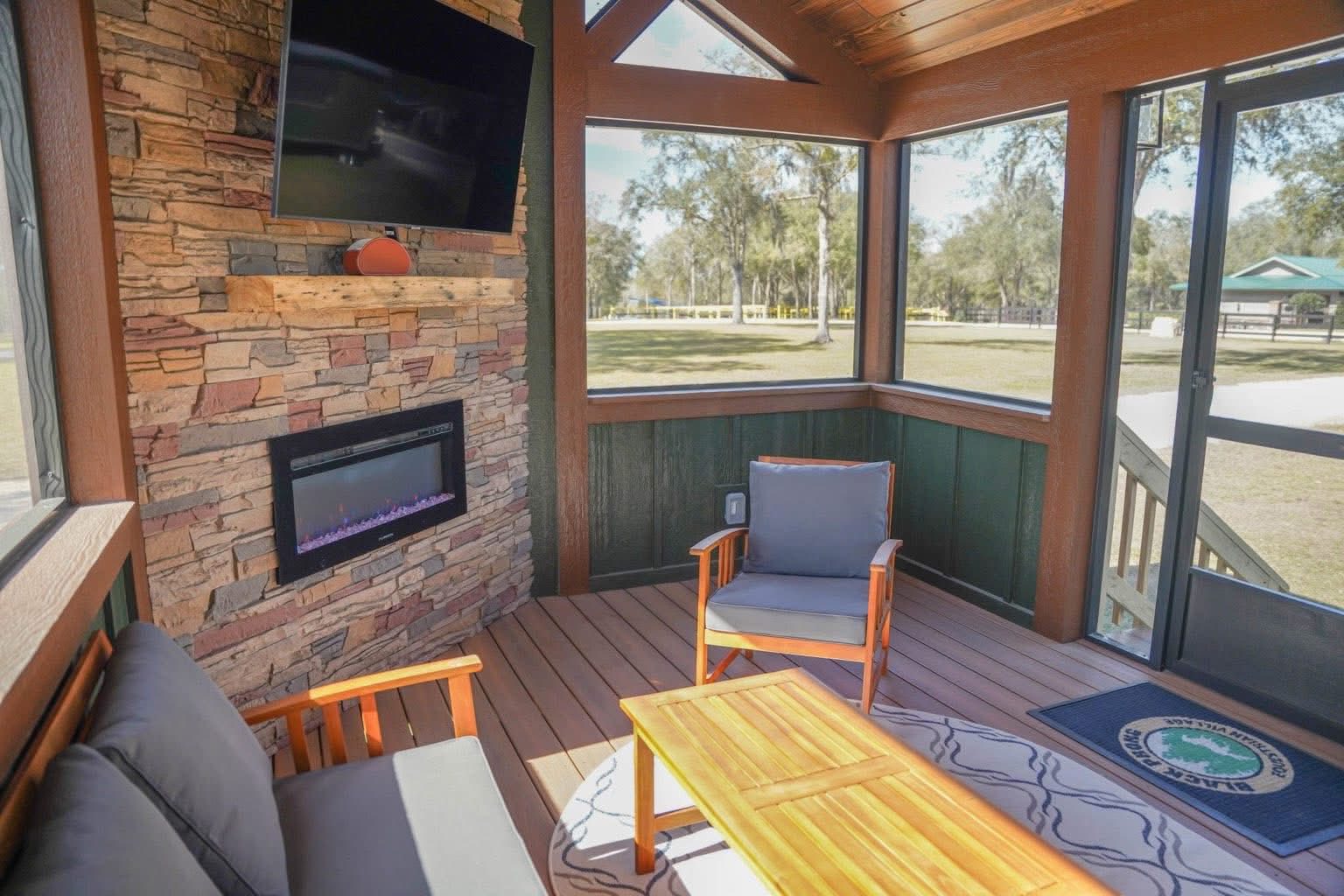 Cozy sunroom with stone fireplace, wall-mounted TV, and outdoor view. Features wooden seating, a table, and large windows, creating a warm, inviting ambiance.