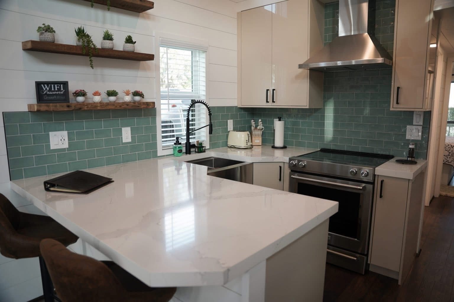 Modern kitchen with teal subway tile backsplash, white countertops, and stainless steel appliances. Open shelves display plants and small decor. Cozy vibe.