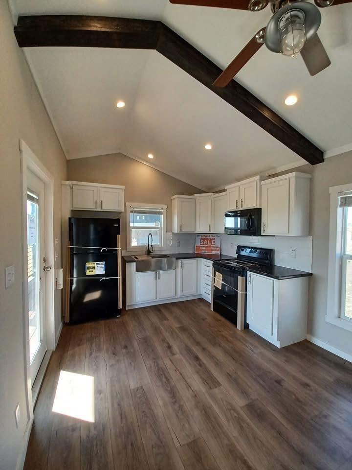 Modern kitchen with vaulted ceiling and wooden beams. Features white cabinets, black appliances, and wood floor. Bright and clean ambiance.