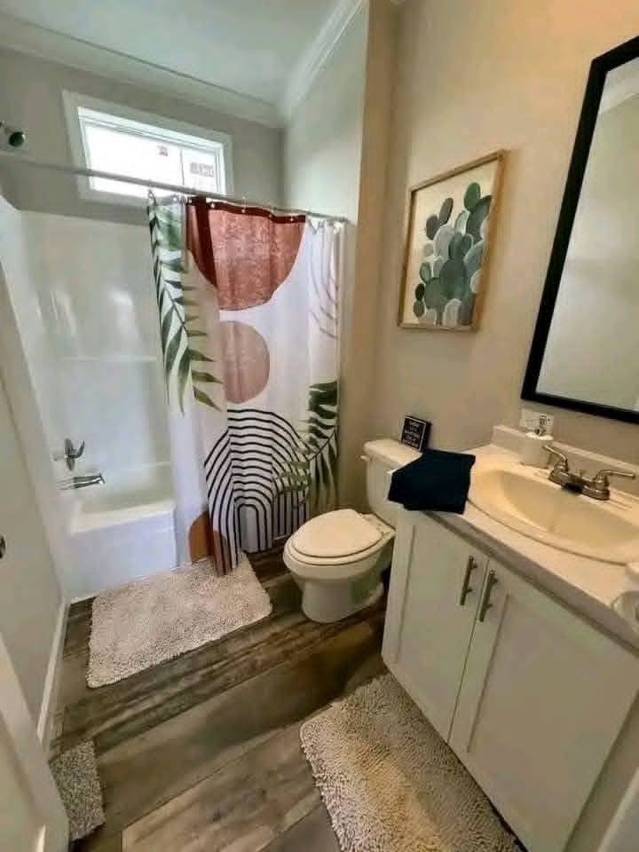 A small, modern bathroom with a bathtub, geometric-patterned shower curtain, wooden floor, two white rugs, sink with cabinet, and framed abstract art.