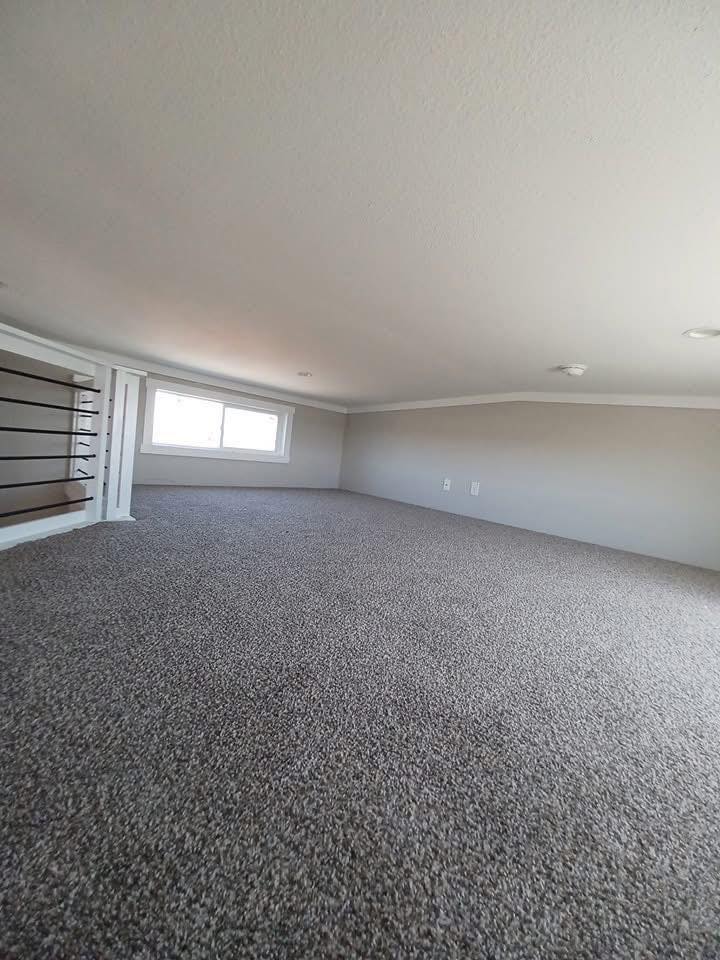 A small, carpeted loft with a sloped ceiling and a low window on the left wall. The neutral tones and soft lighting create a cozy, minimalist feel.