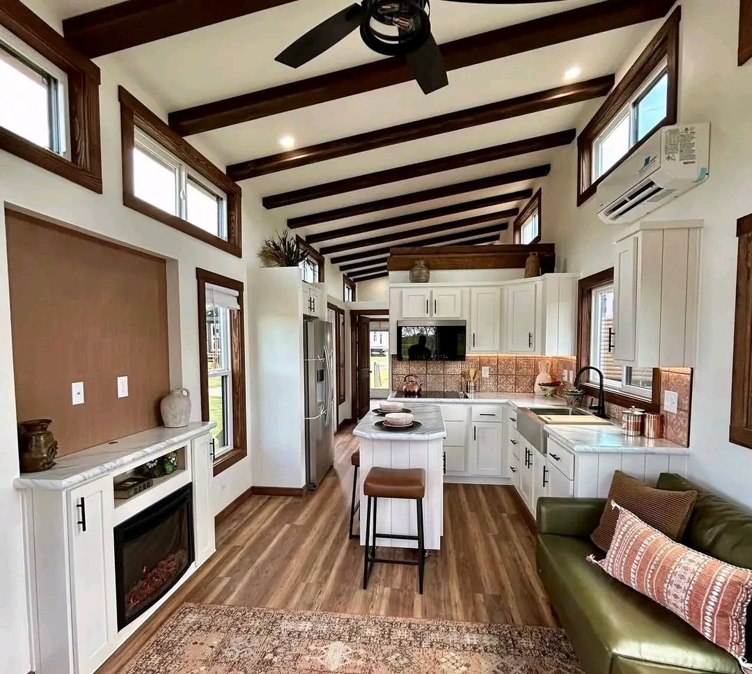 Cozy tiny home interior with wood beams, white kitchen cabinets, and an island. A green couch with pillows sits next to an electric fireplace. Warm and inviting.