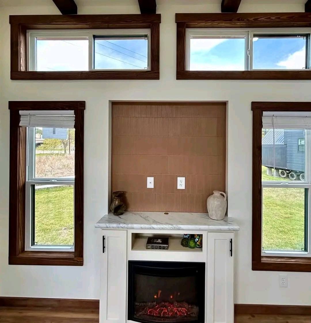 A cozy living room features a small fireplace with red flames, framed by white mantel and a marble top. Large windows with wooden trim allow natural light.