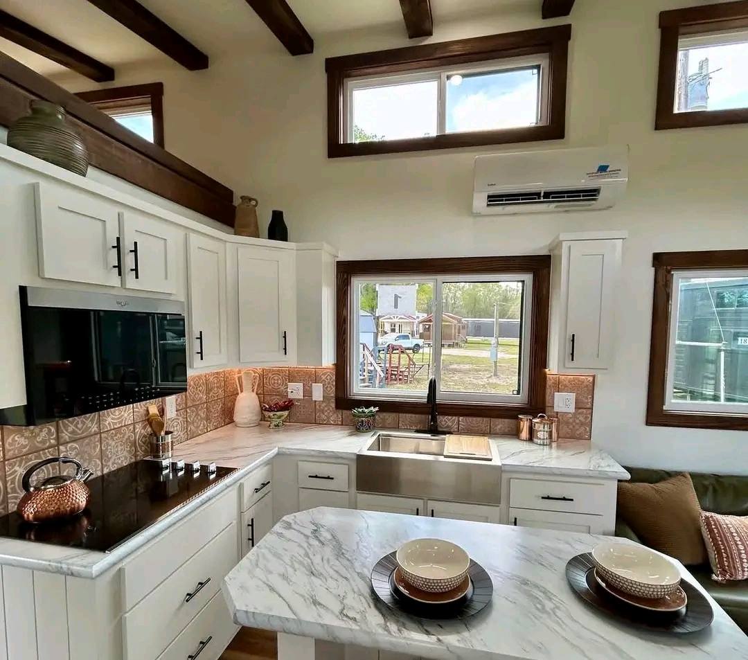 Modern kitchen with white cabinets, marble island, and copper accents. Large windows offer a view of an outdoor play area, creating a bright, airy feel.