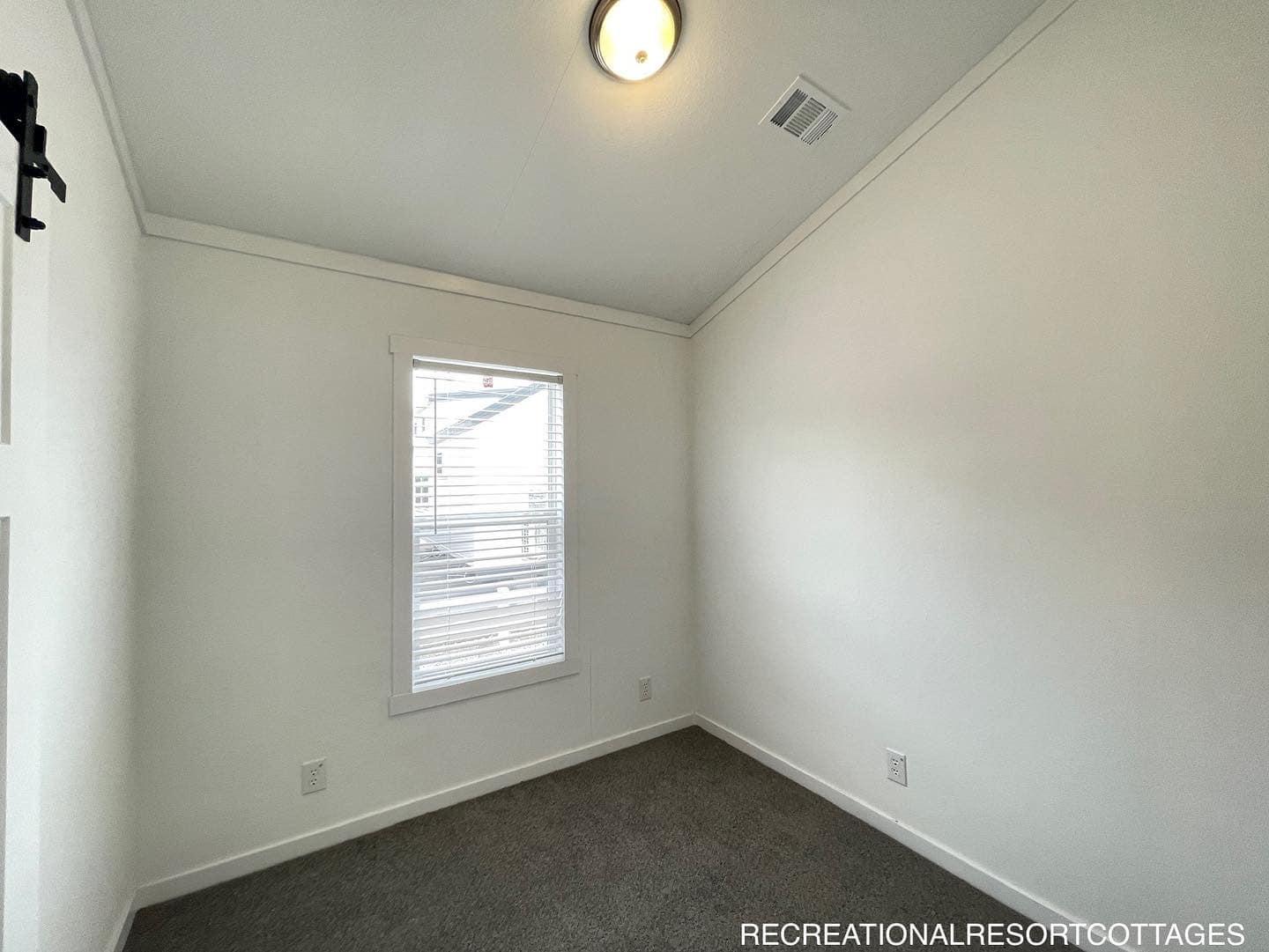 Simple, small empty room with white walls, dark carpet, and a single window with blinds. Ceiling light fixture and a visible air vent.