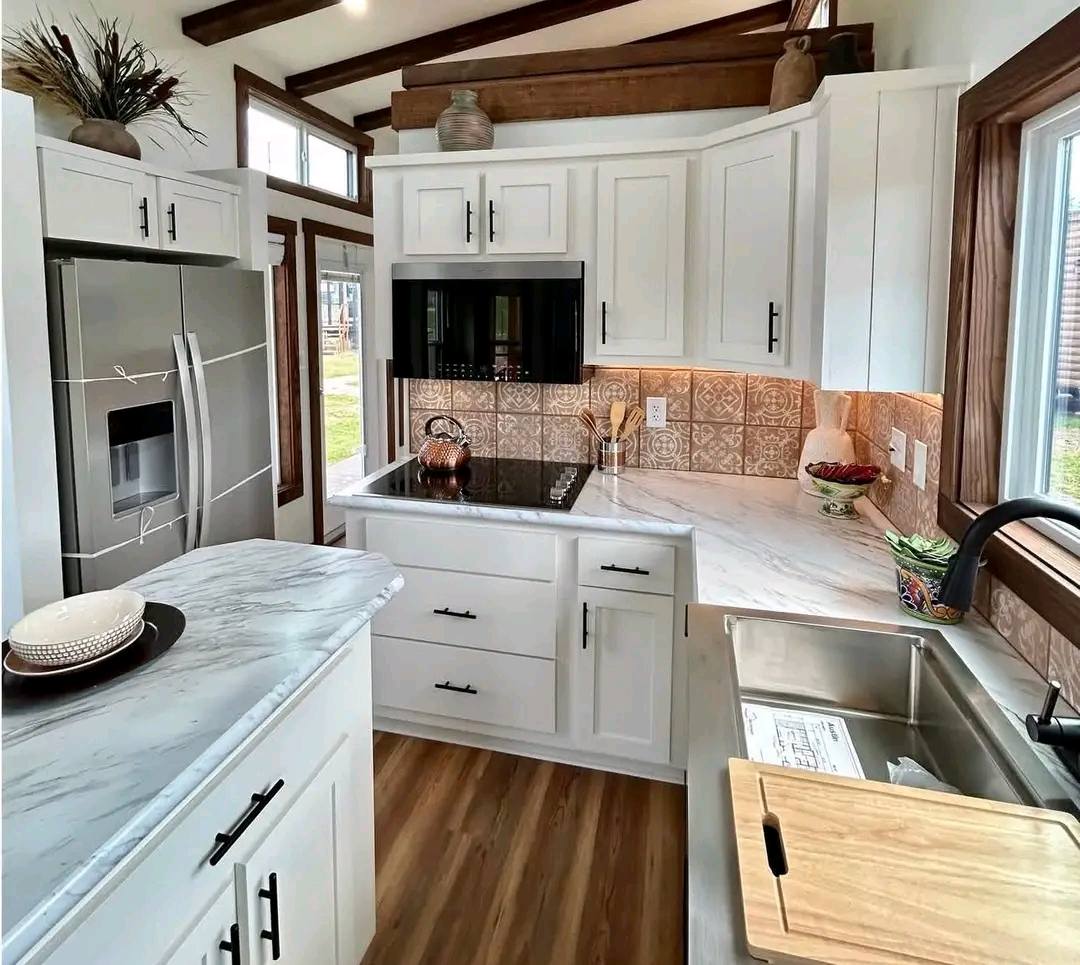 A small, cozy kitchen with white cabinets and marble countertops. It features a stainless steel fridge, a black cooktop, and warm wood accents.
