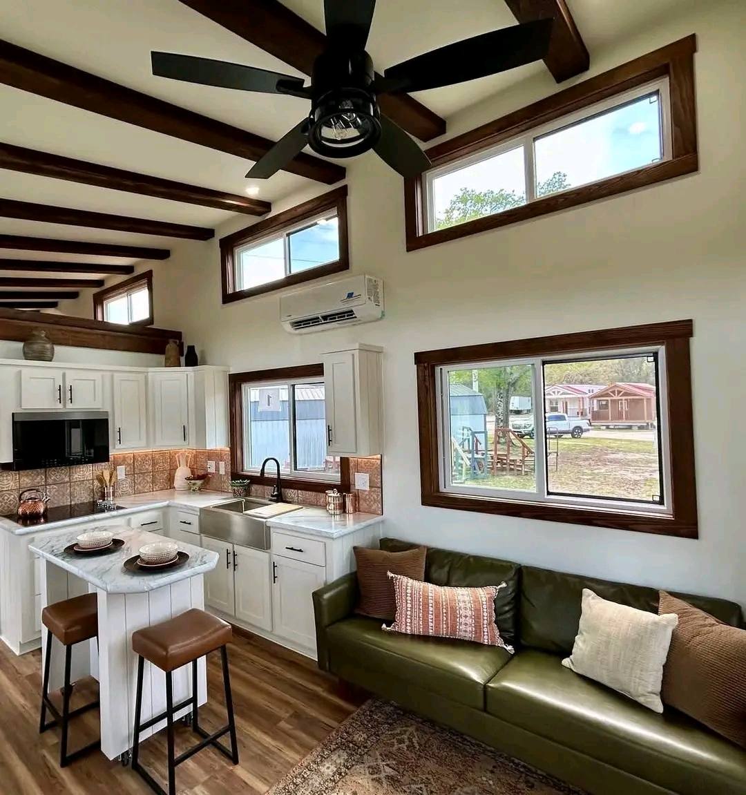 Compact modern kitchen and living area with white cabinets, an island, green sofa, and bar stools. Bright windows and wooden beams enhance coziness.