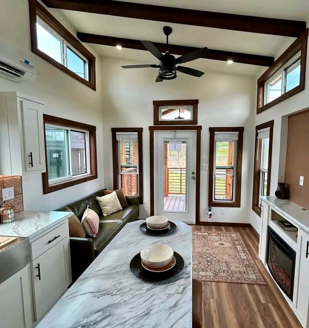 Modern tiny house interior with exposed beams, white walls, and large windows. Central marble countertop with dishes, cozy sofa, and electric fireplace.