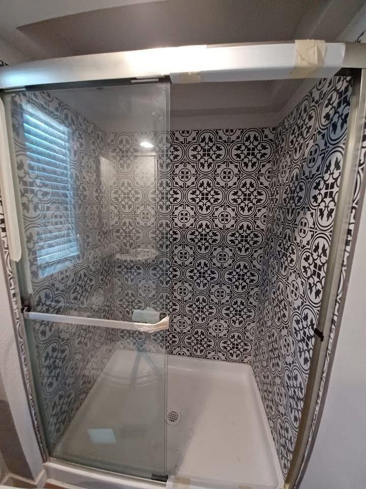 Shower with a sliding glass door, featuring intricate black and white mosaic tiles. The modern design is illuminated by natural light from a nearby window.