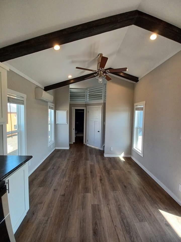 Cozy, narrow room with wood floors, vaulted ceiling, and dark beams. A ceiling fan, bright natural light from windows, and white walls create an airy feel.