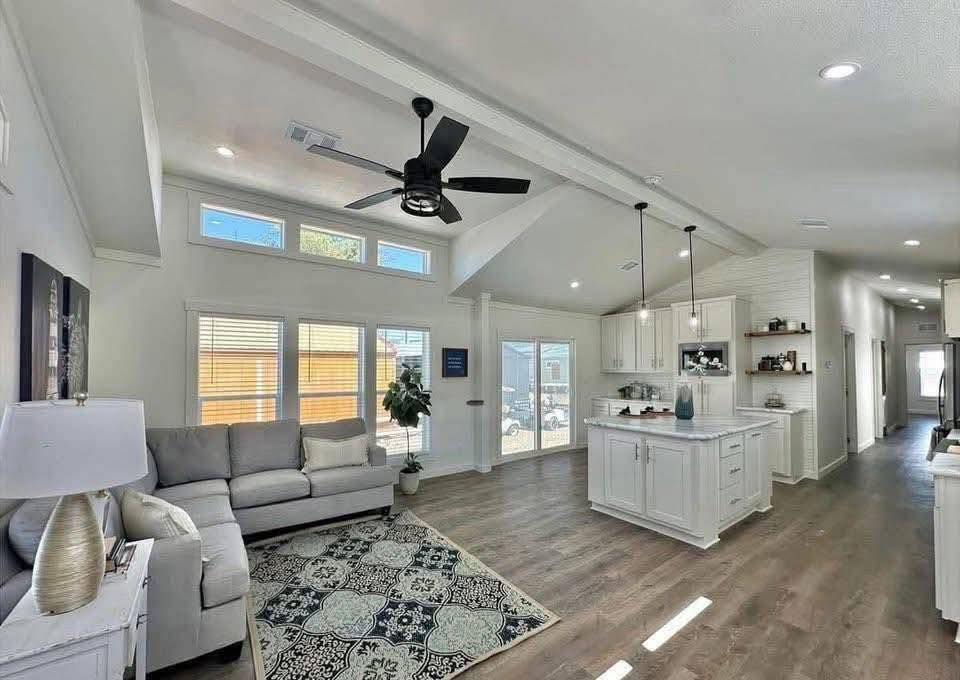 Spacious living room and kitchen with vaulted ceiling, neutral tones, and modern decor. Features a ceiling fan, white cabinets, and natural light. Cozy and inviting.
